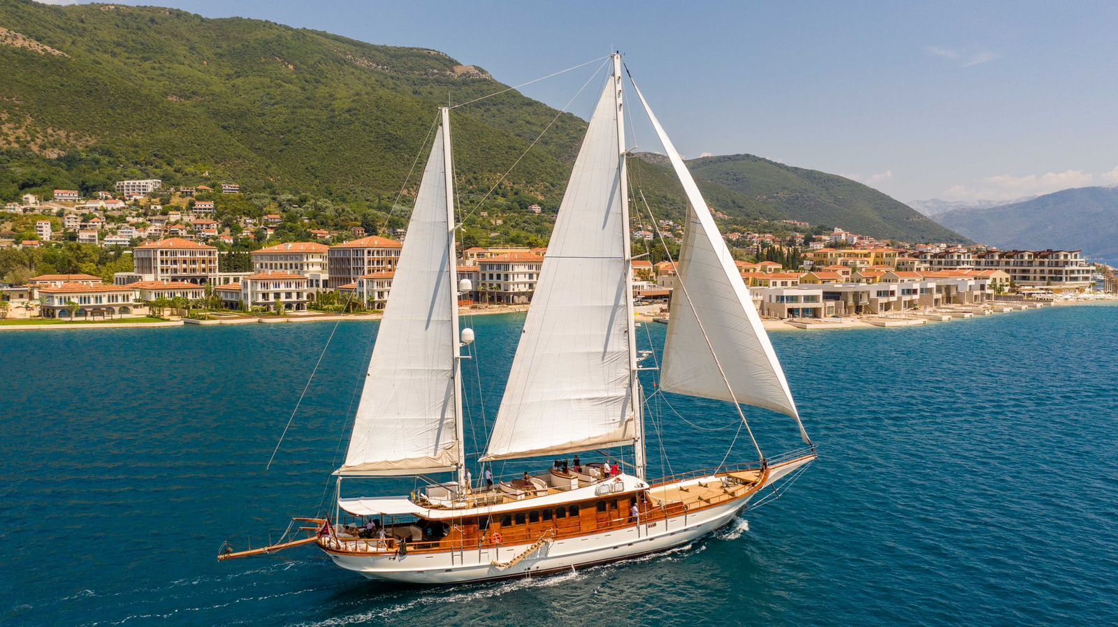 Sailboat with white sails cruising on blue water near a coastal town and green hills