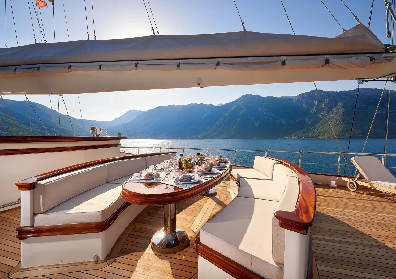 Sunlit yacht deck with white lounge seating, a table, and mountains across a calm lake