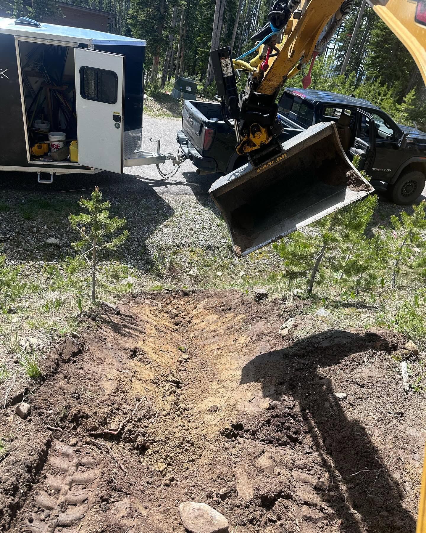Excavator bucket and truck beside a muddy dirt path in a wooded area.
