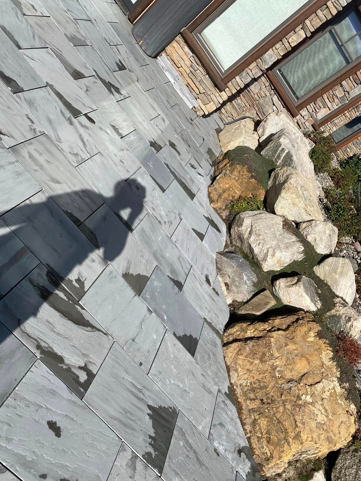 Stone walkway beside rocks and a brick house, with a person’s shadow on the slate paving.