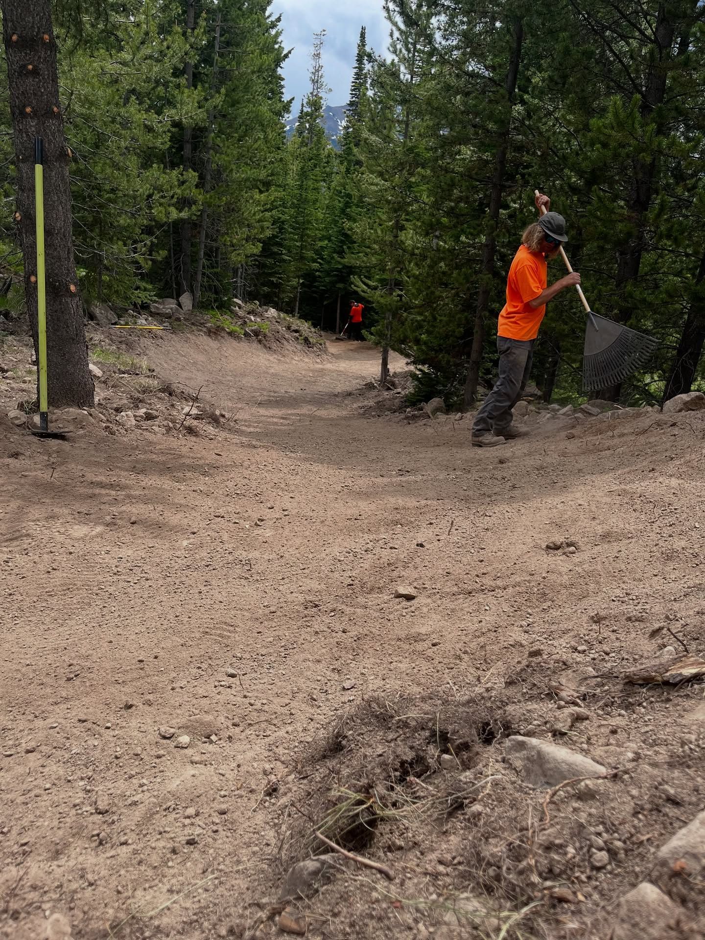 Mountain biker in orange jacket navigating a dusty forest trail with another rider ahead