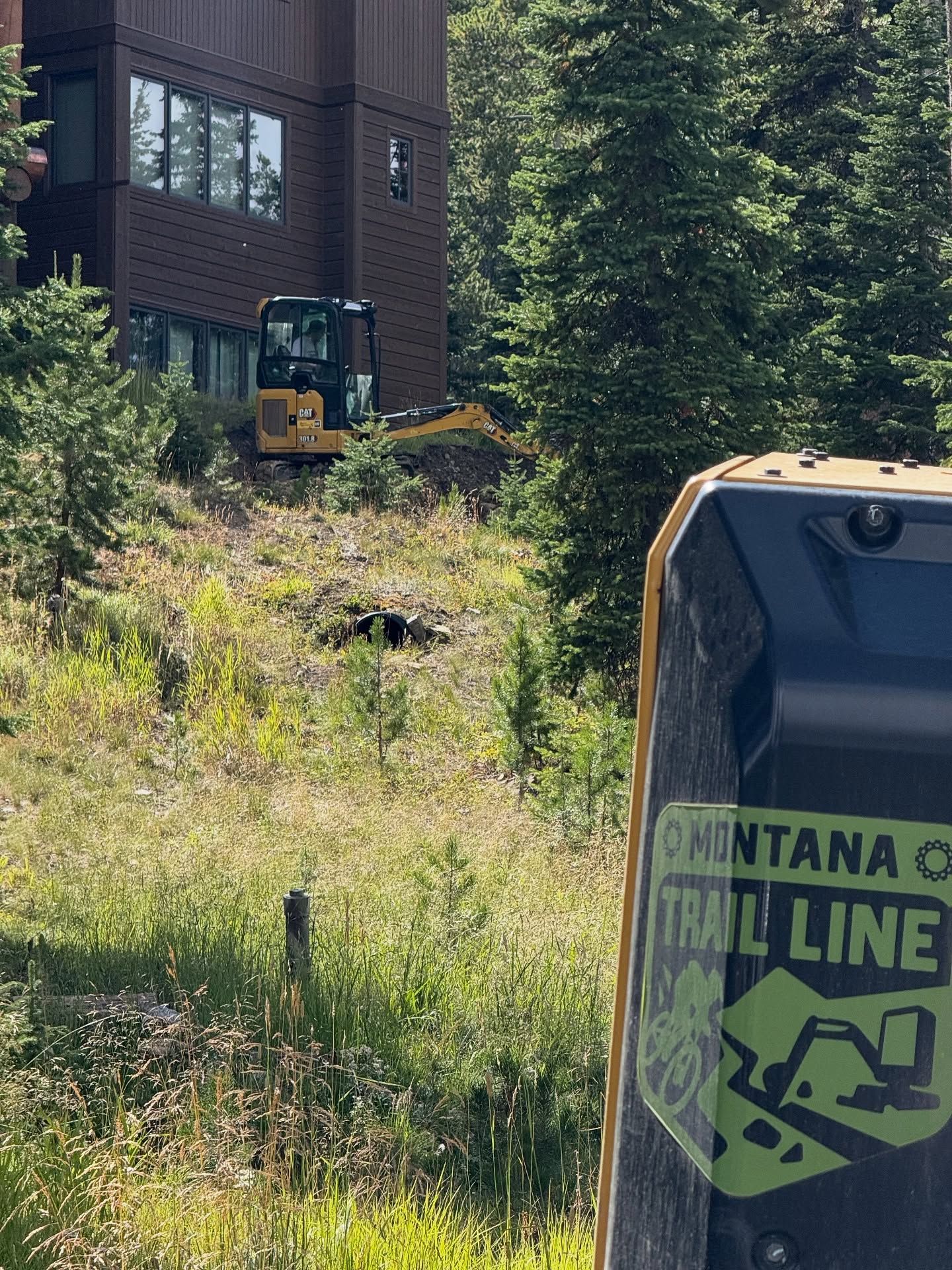 Montana City Trail Line sign beside a grassy hillside and brick building near a forest