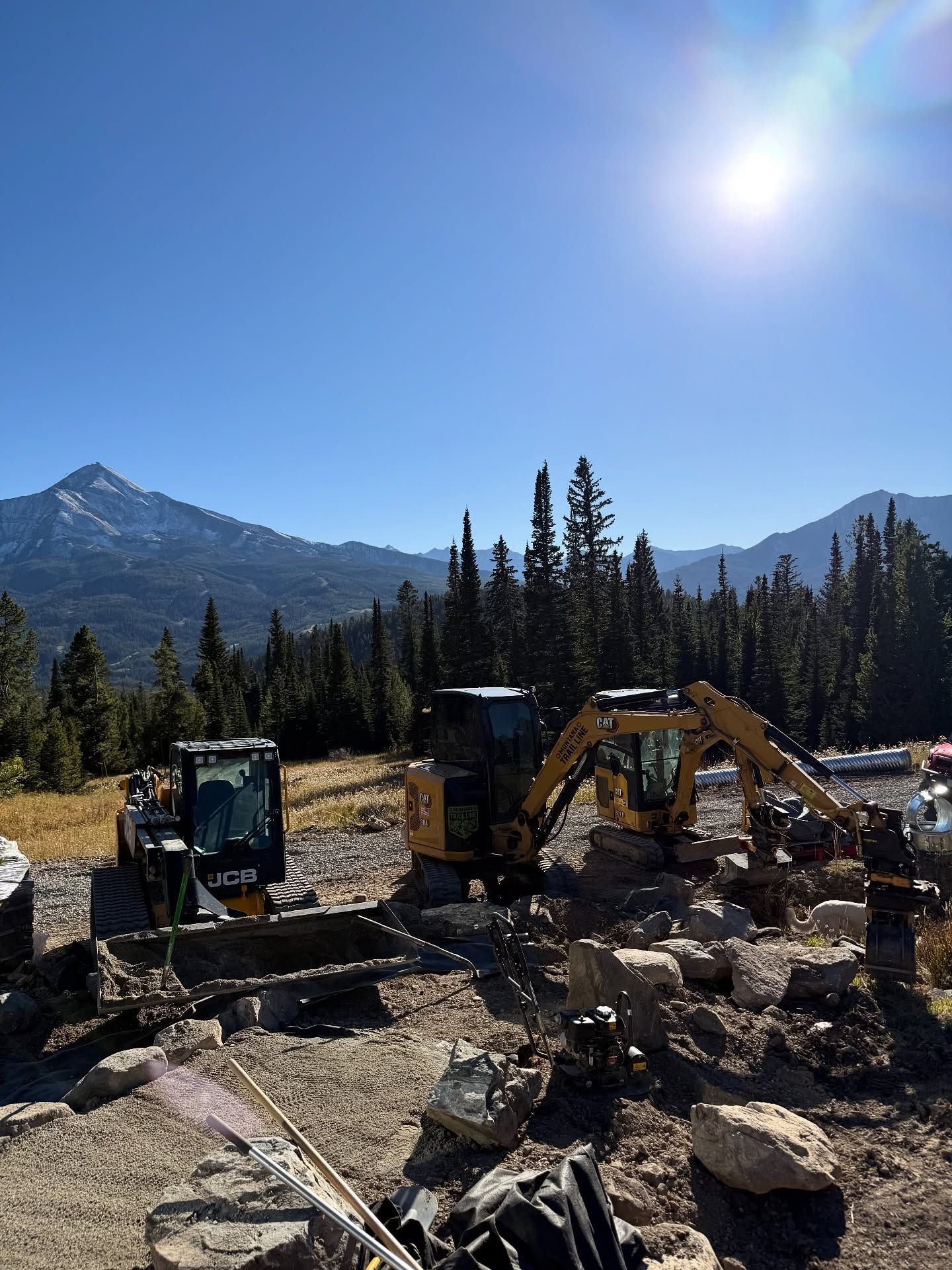 Construction excavators clearing rocks in a mountain meadow under a bright sun.
