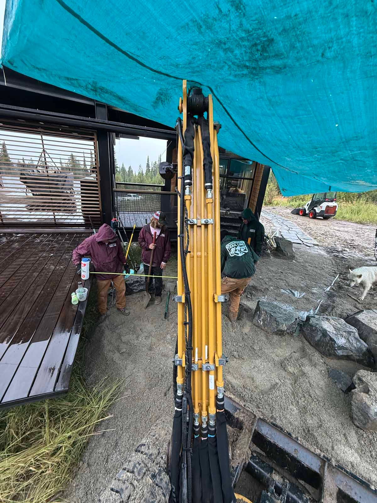 Yellow excavator at a muddy construction site under a blue tarp, with two workers nearby.