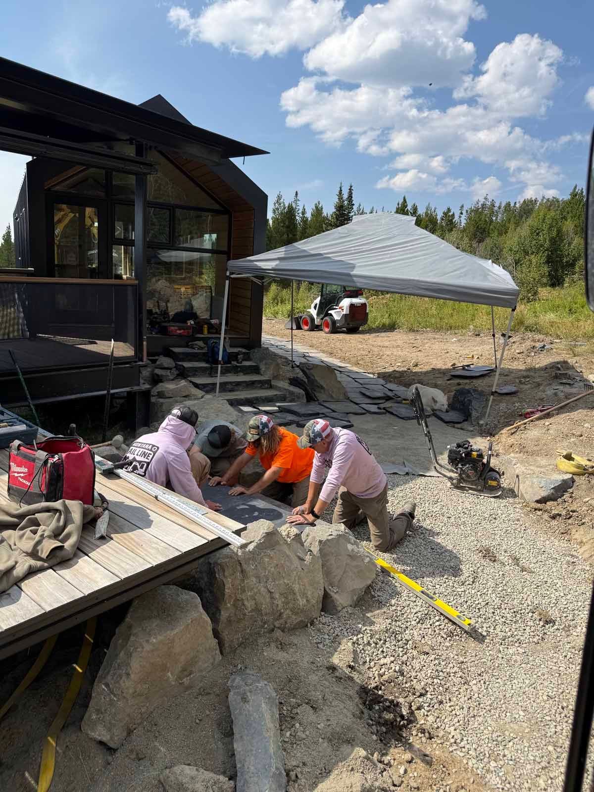 Workers laying gravel beside a modern house under construction, with a canopy tent and cloudy sky.