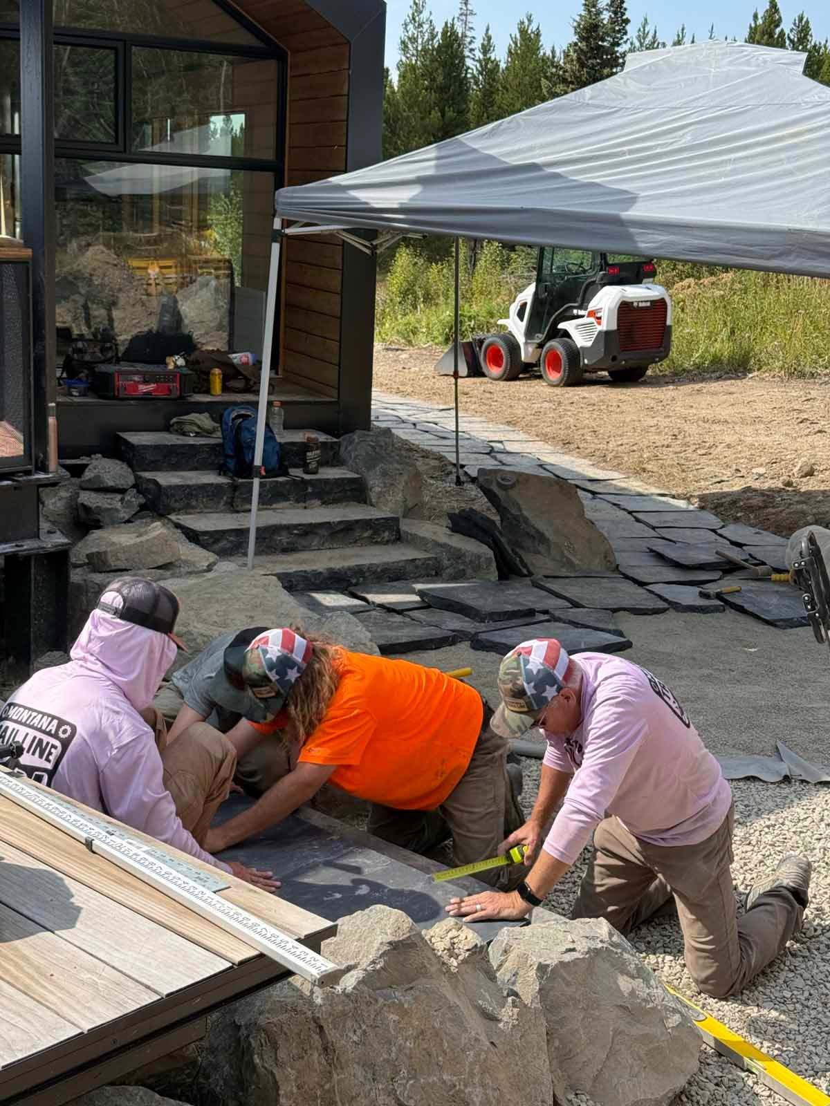 Three workers in safety vests install stones beside a house under a white canopy.