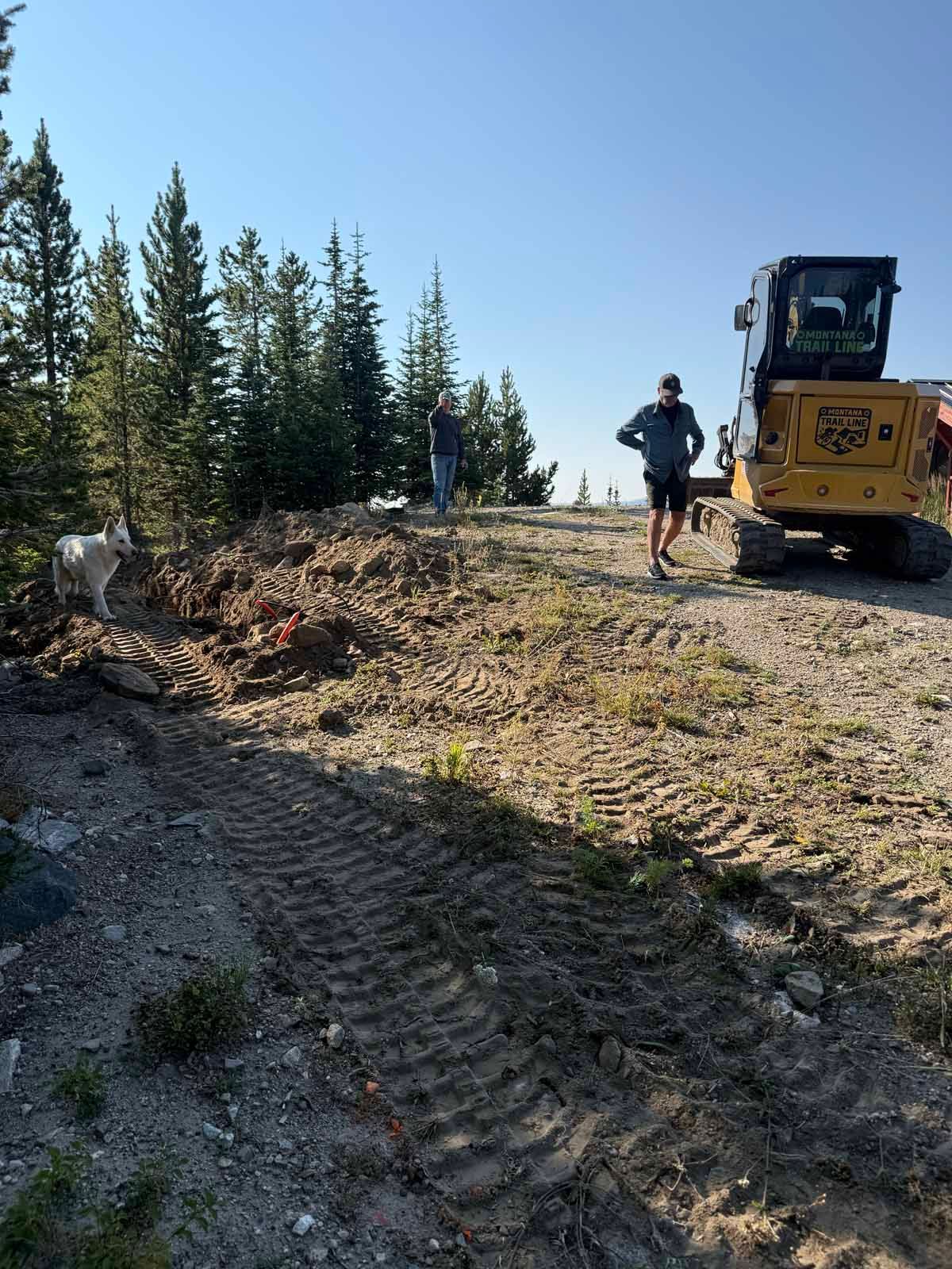 Construction worker beside yellow excavator on a dirt road in a forest clearing.