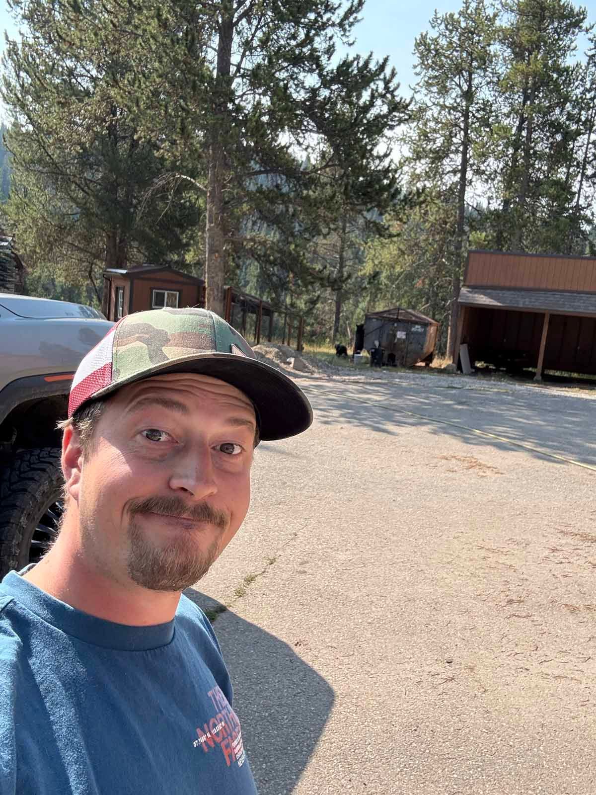 Selfie of a smiling man in a cap at a wooded campsite with cabins and parked vehicles
