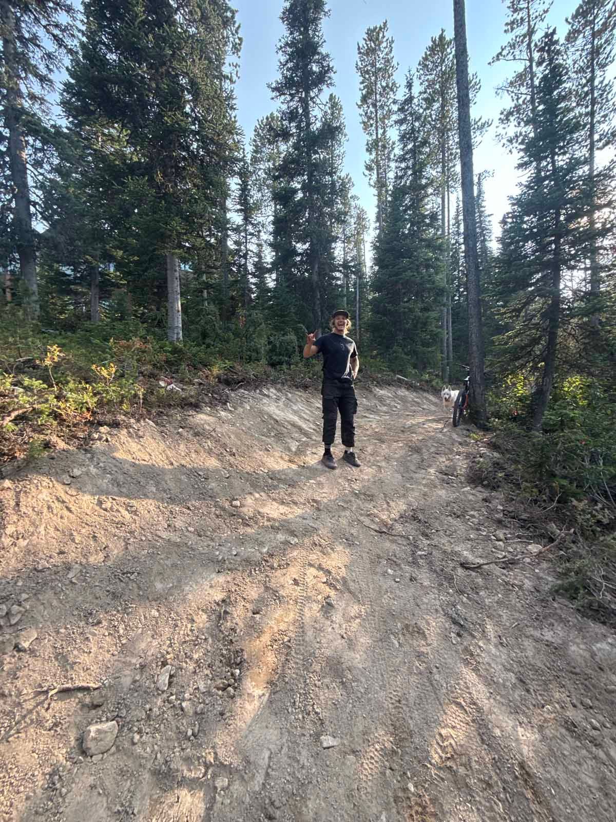 Person standing on a dirt trail in a forest of tall pine trees.