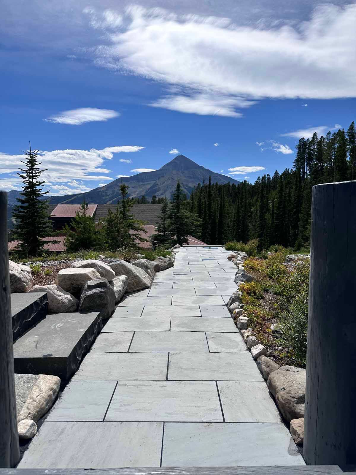 Stone walkway leading toward a mountain under a blue sky, with trees and rocks on both sides