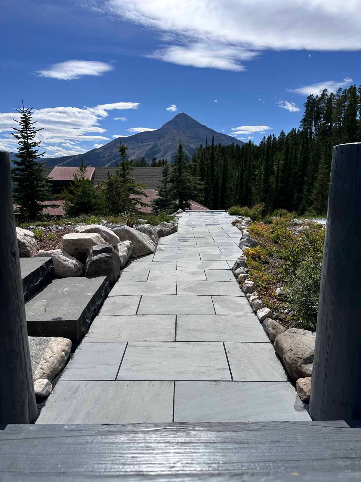 Stone walkway through a mountain garden with rocks, trees, and a peak under a blue sky.