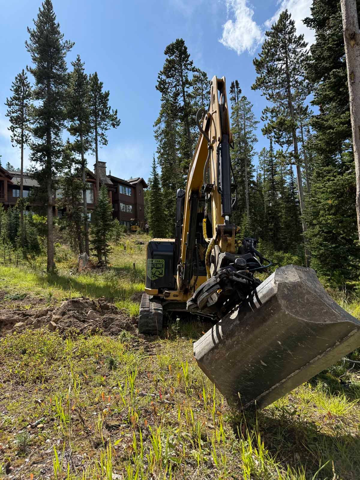 Excavator working on a grassy hillside near pine trees and cabin buildings under a blue sky.