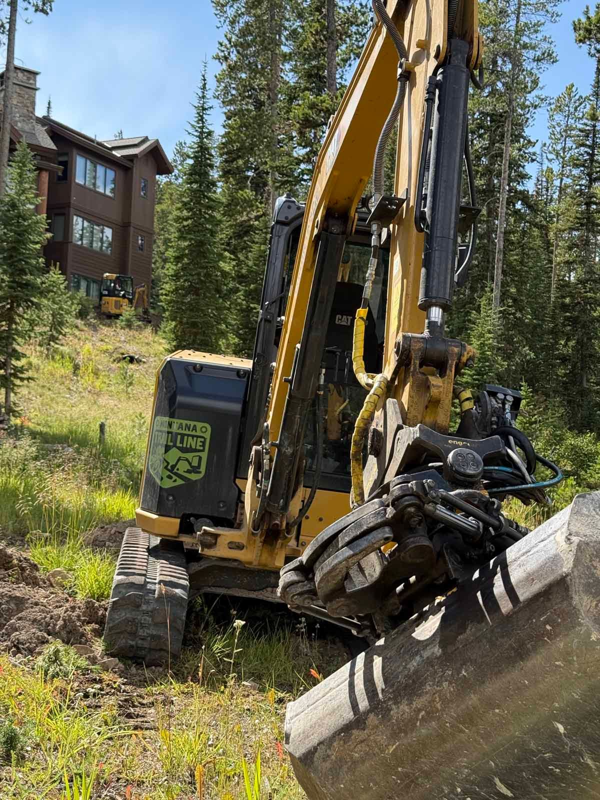 Yellow excavator clearing a hillside beside a building and tall pine trees