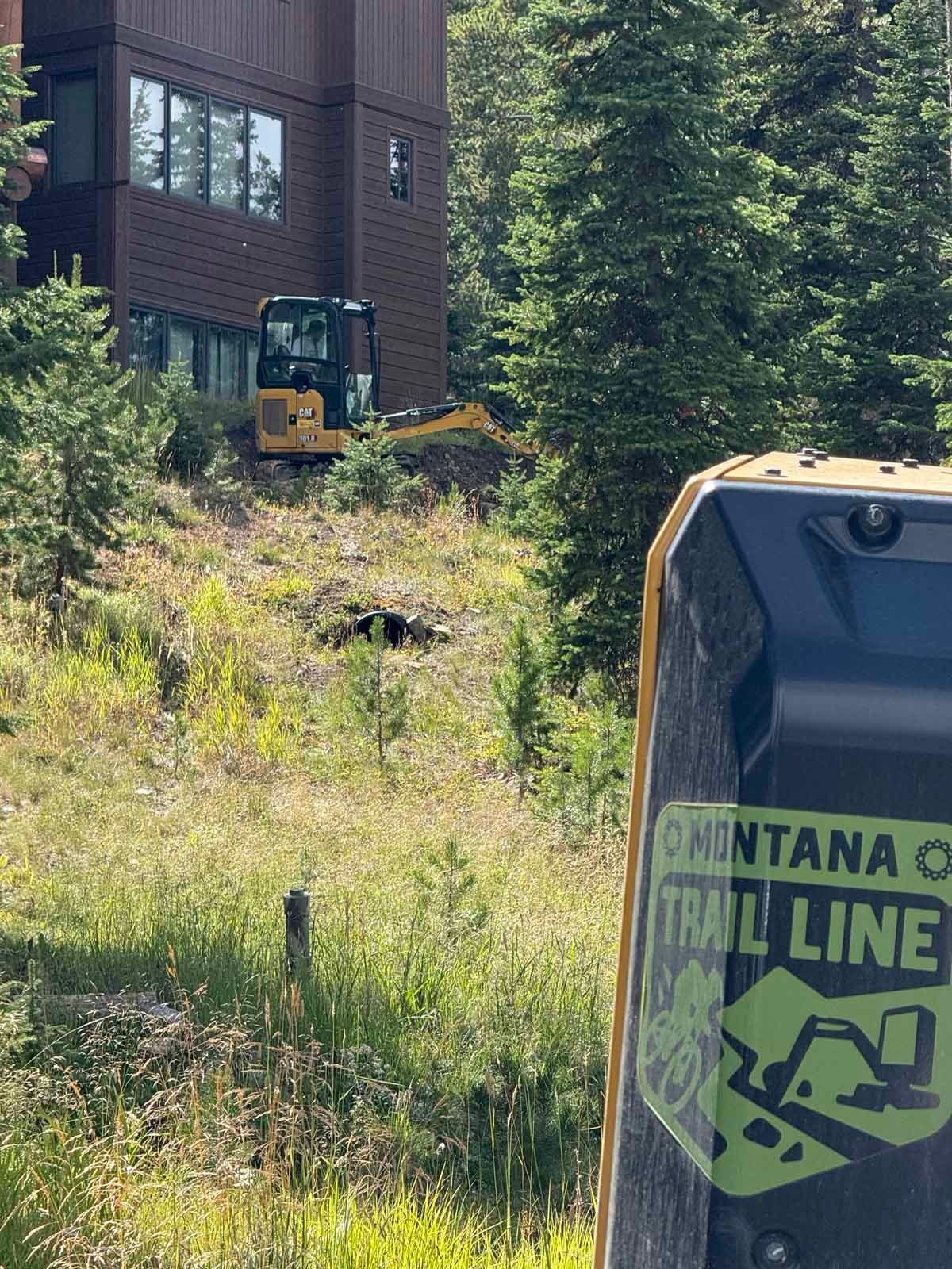 Orange excavator beside a brick building in a wooded, grassy area, with a Montana Trail Line sign in the foreground