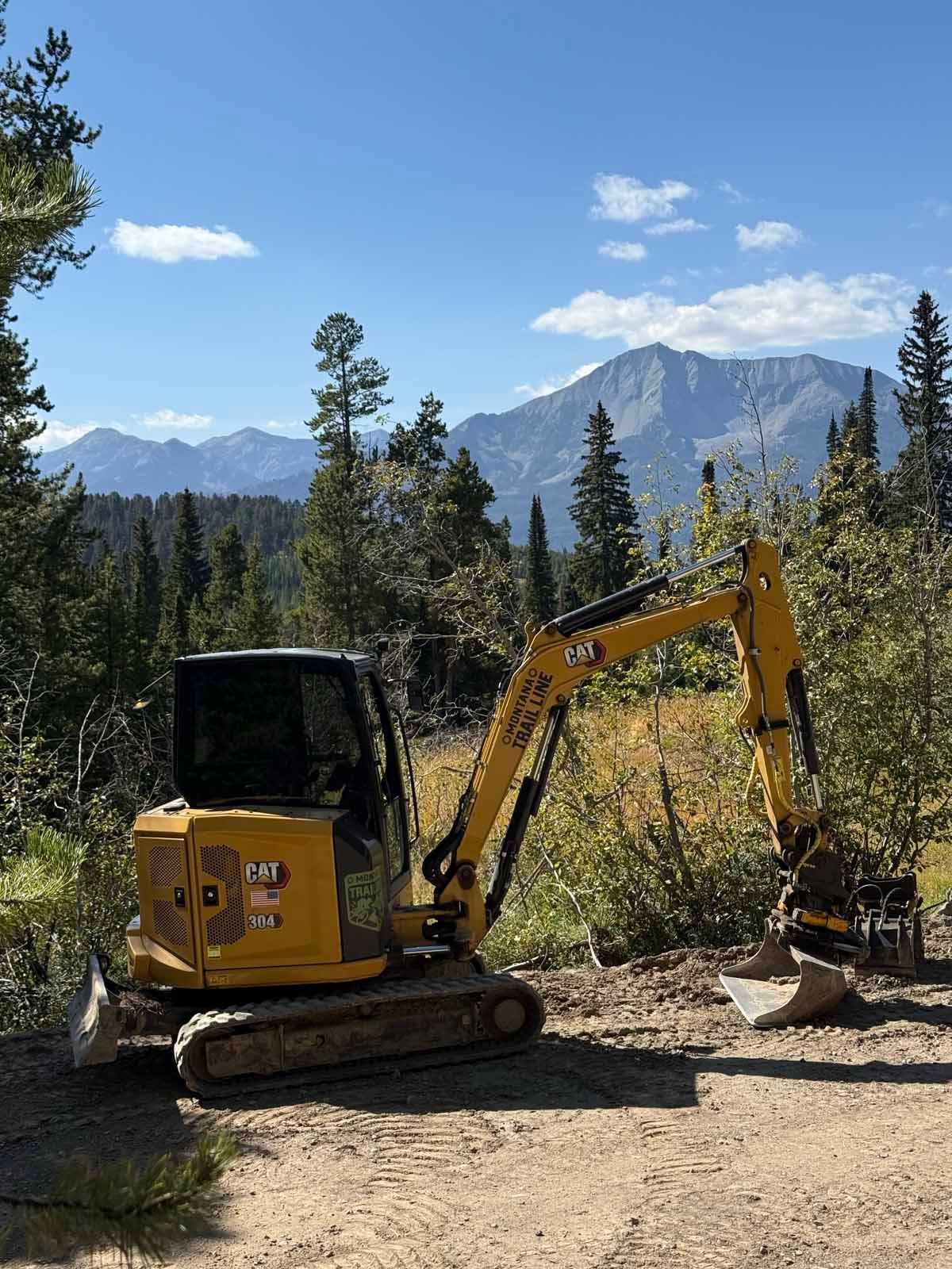 Yellow excavator working on a dirt path in a forested mountain landscape under a blue sky