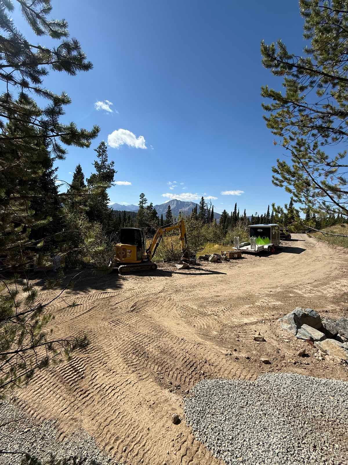 Mountain overlook with dirt road, tall pines, and distant snowy peaks under a clear blue sky