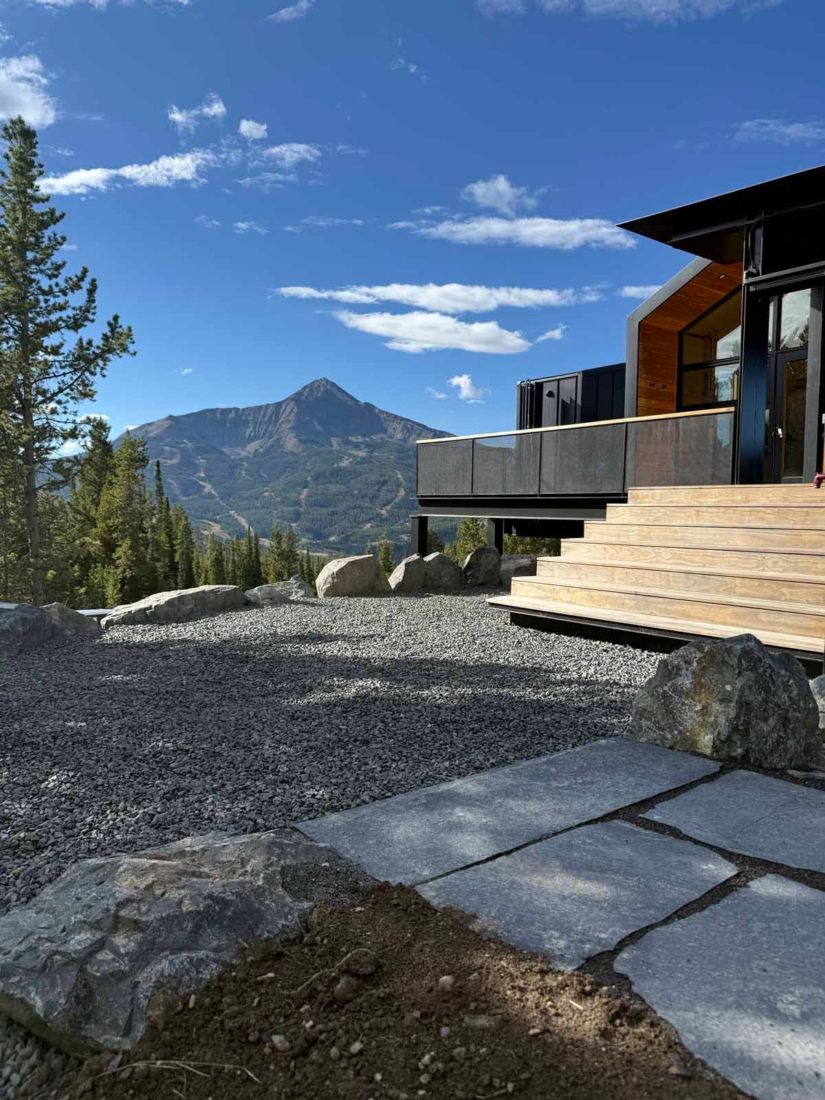 Modern mountain home with stone patio, rocky yard, and pine trees overlooking a distant mountain.