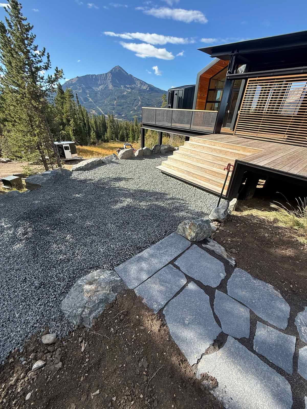Mountain cabin deck beside gravel path with stone steps, pine trees, and a distant rocky peak under blue sky