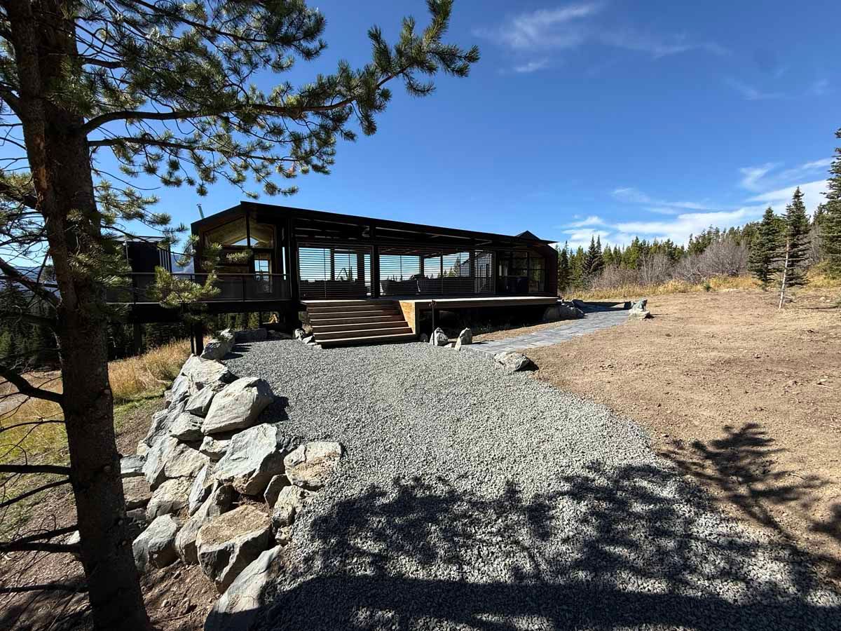 Wooden cabin with deck in a rocky, dry clearing beside pine trees under a blue sky