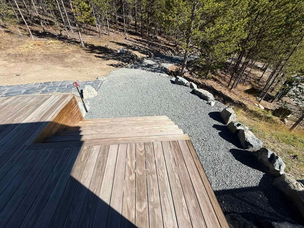 Wooden deck overlooking a gravel yard and wooded hillside in bright sunlight