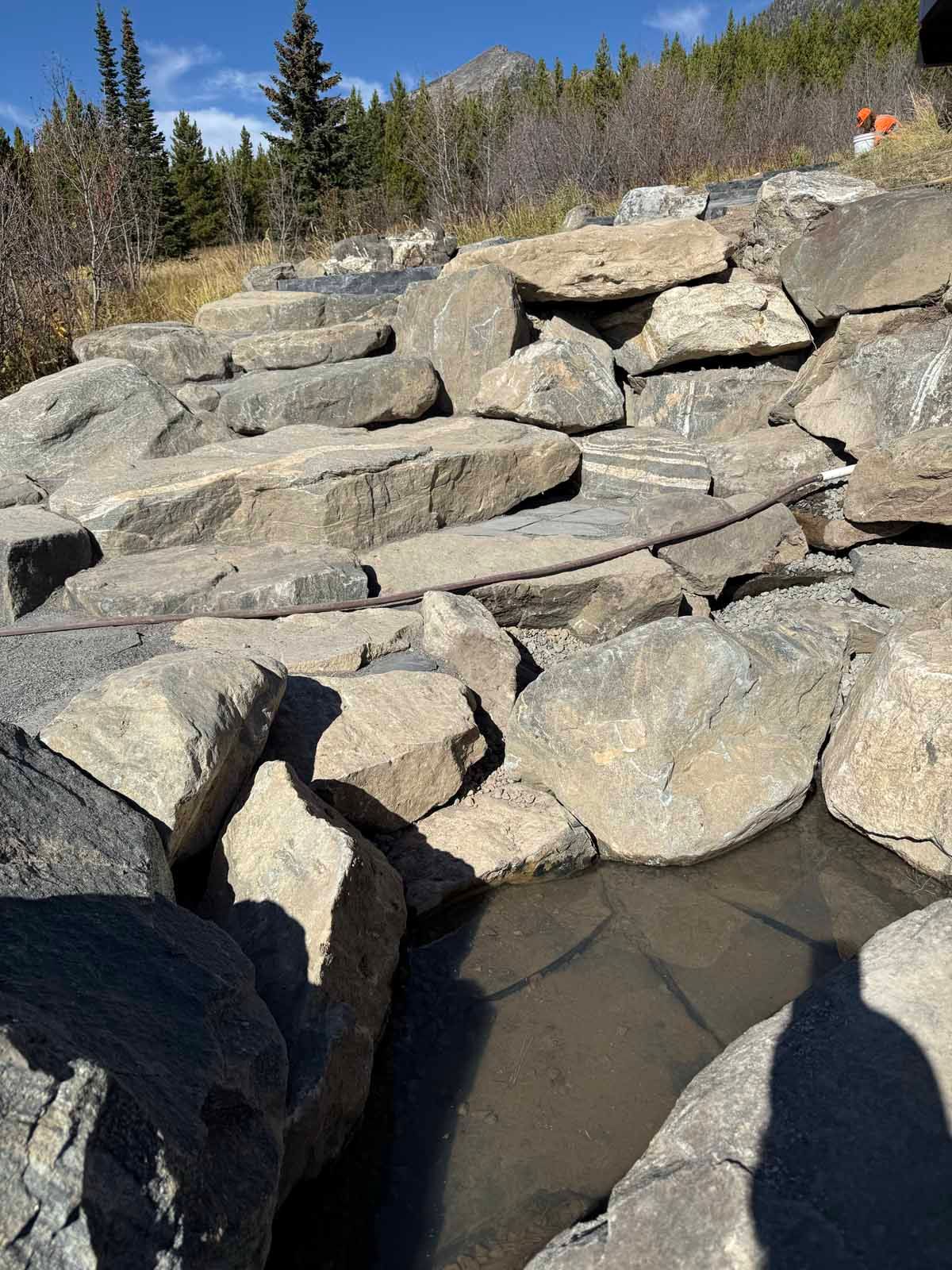 Stacked stone retaining wall beside muddy water under a clear blue sky in a wooded area