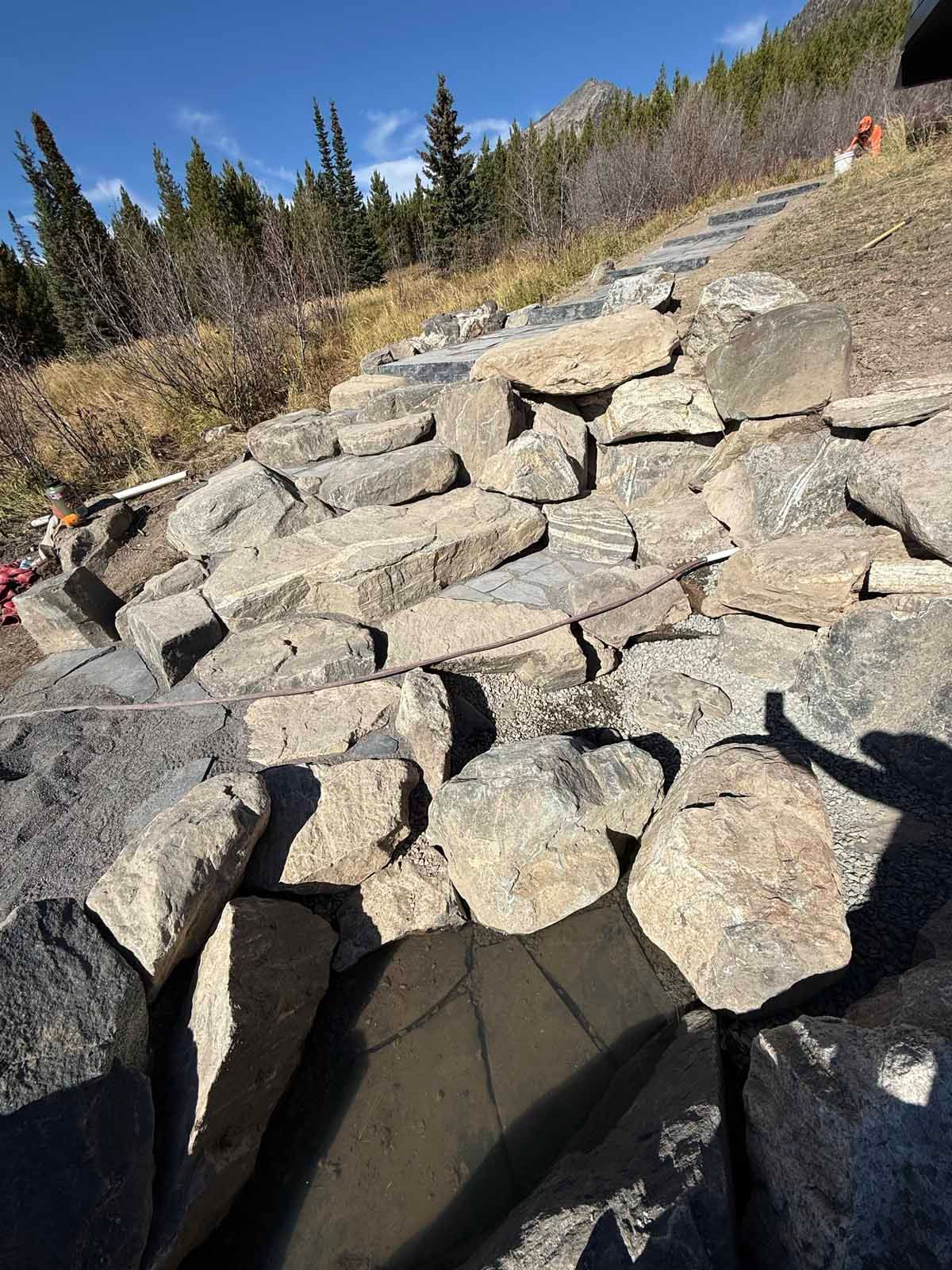 Rocky hillside trail with large gray boulders under a clear blue sky and pine trees.