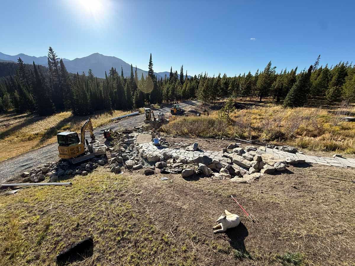 Rock-lined fire pit in a grassy mountain clearing with pine trees and distant peaks under a blue sky