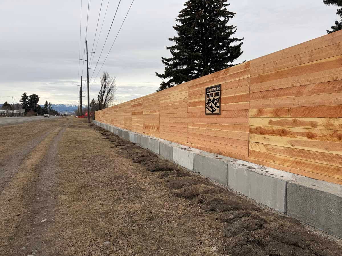 Wooden privacy fence along a gravel roadside with concrete base and a tall evergreen tree behind it