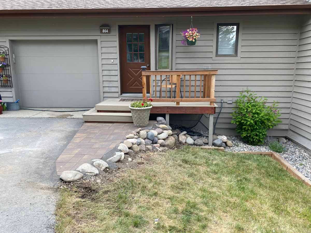 Front entrance of a house with a wooden porch, stone-lined path, garage, and small front lawn