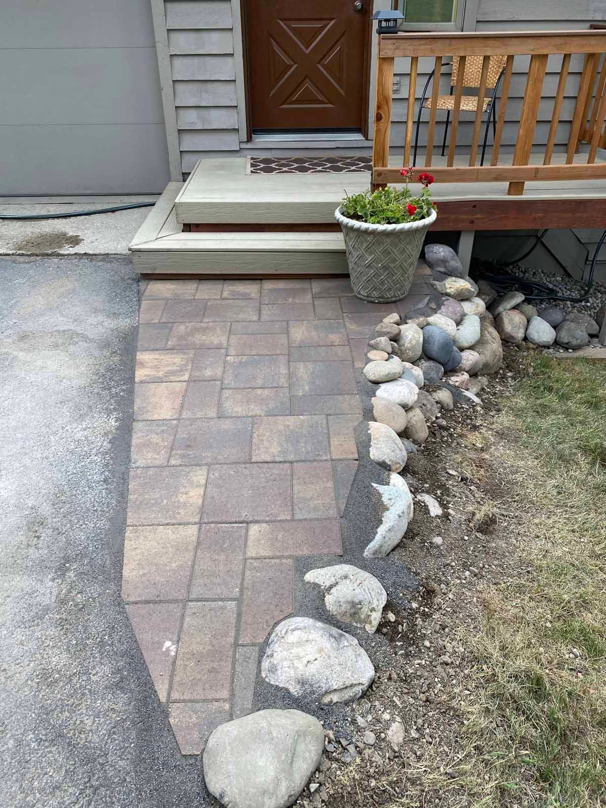 Brick-paver walkway beside a house, lined with river rocks and leading to a front porch.