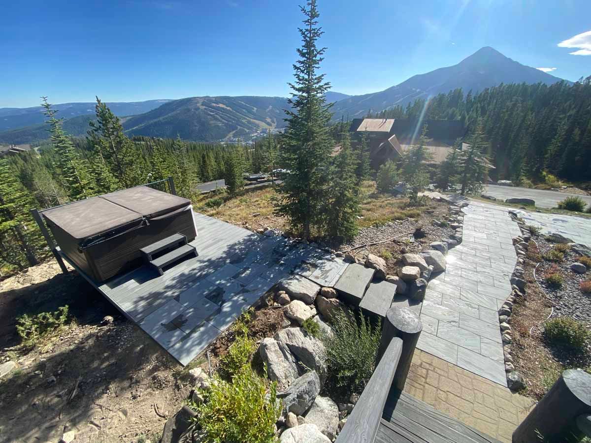 Mountain patio with benches, stone path, pine trees, and distant peaks under a clear sky