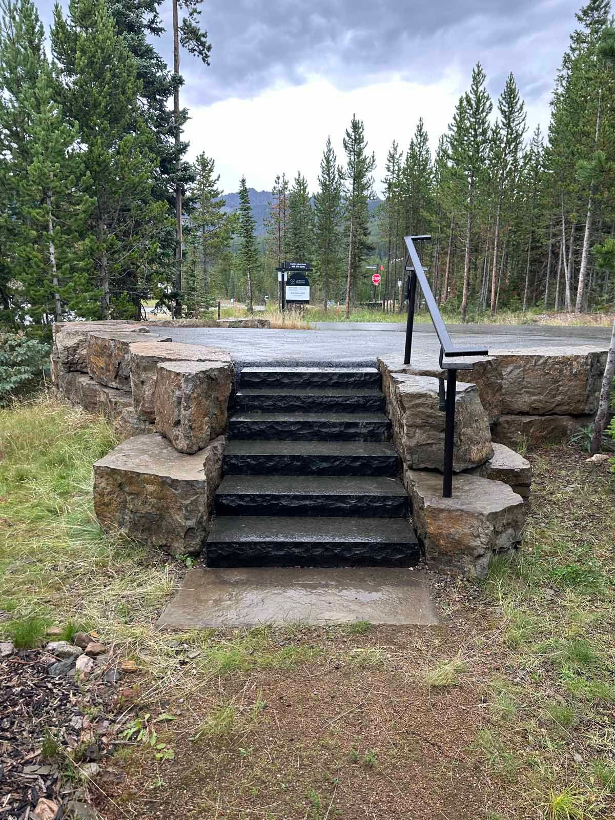 Stone steps leading to a gravel path in a forested park, with a metal handrail and cloudy sky.