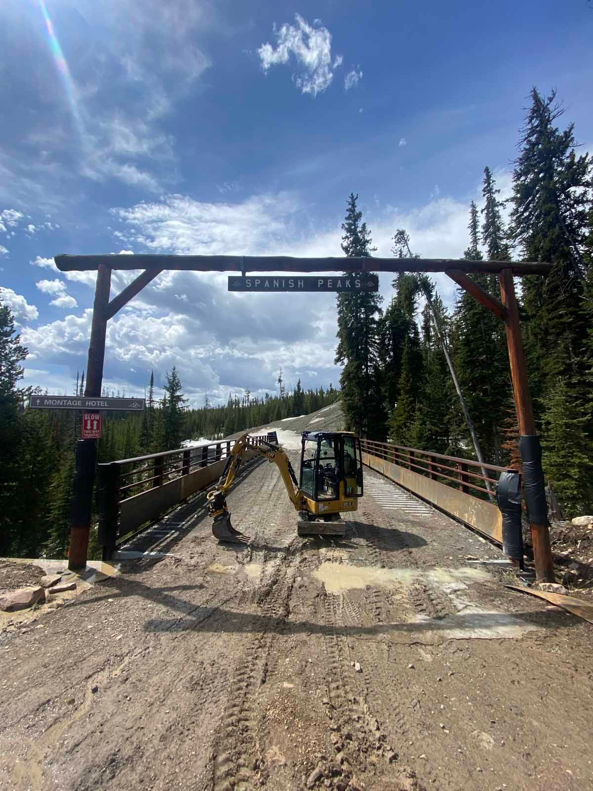 Off-road construction vehicle under a wooden entrance arch on a muddy forest road under blue sky