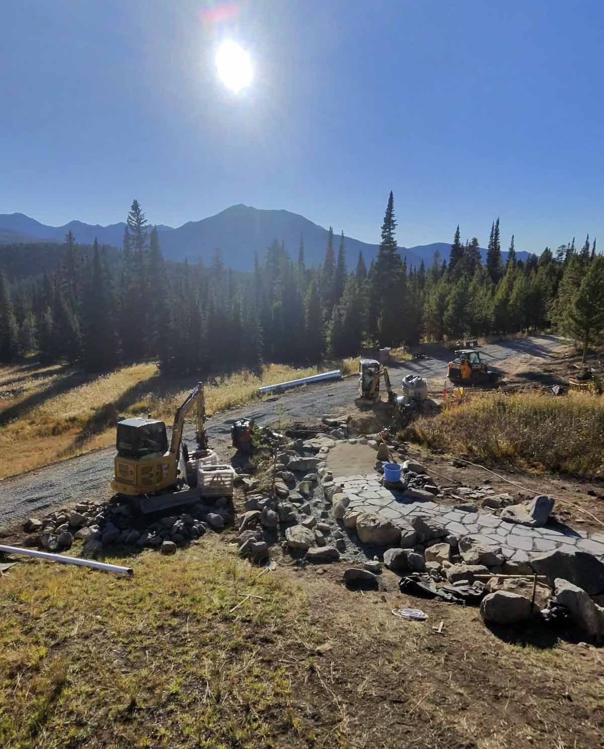 Sunlit mountain landscape with a winding stone path, pine trees, and distant peaks under a clear blue sky