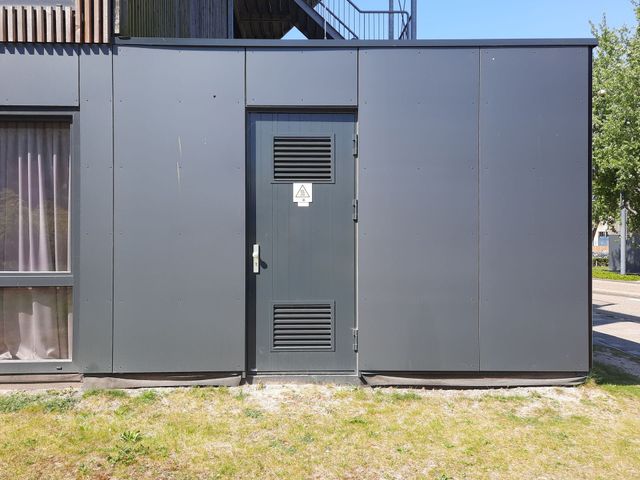 Gray utility building with a central metal door and vents, viewed from outside on a grassy lot.