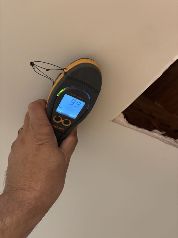 Hand holding a stud finder near a hole in a white ceiling, the display shows 