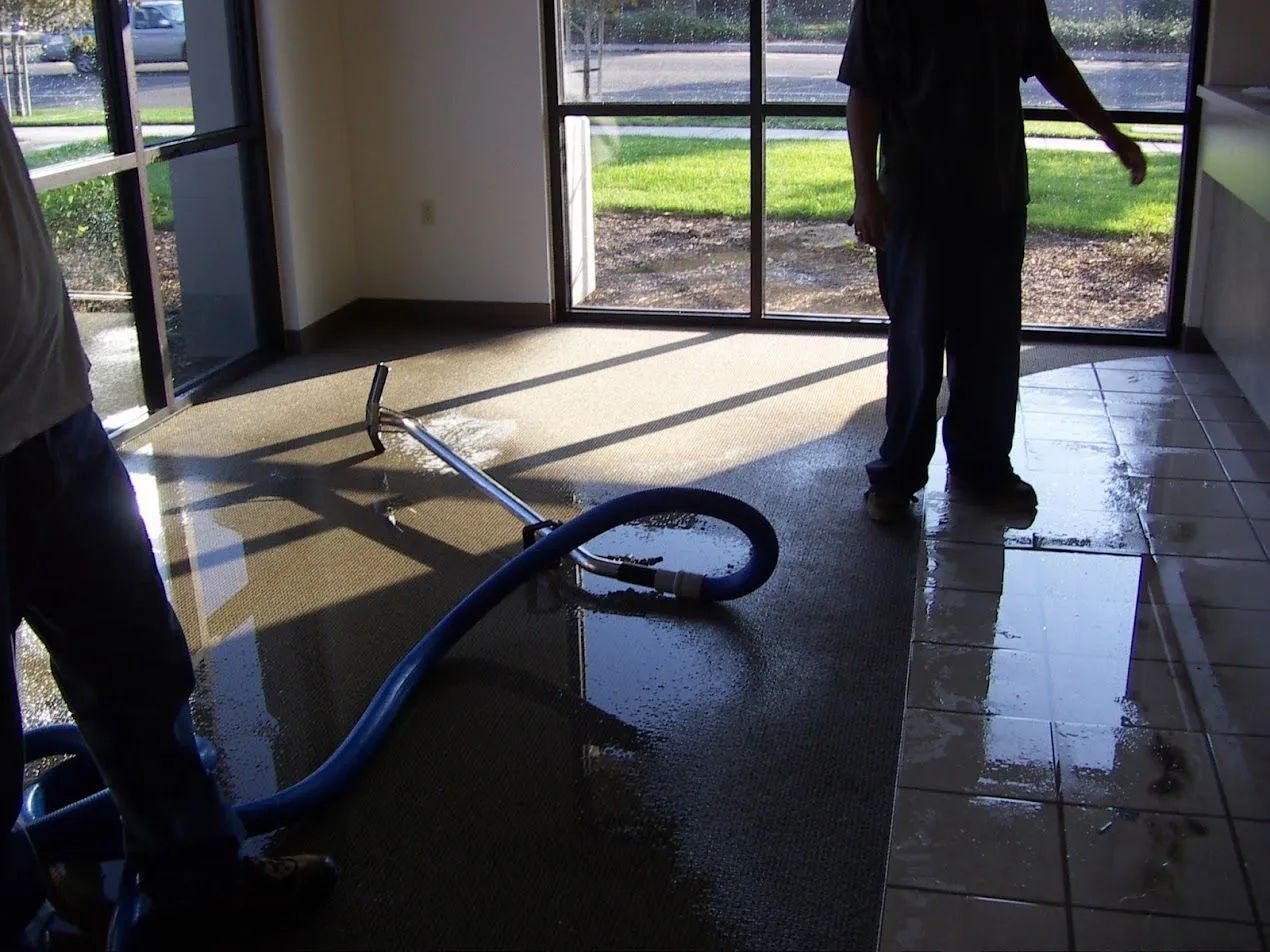 Flooded room being cleaned with a vacuum hose, sunlight through glass doors.