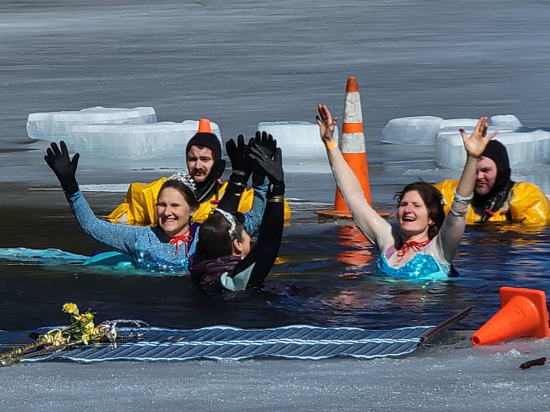 Image shows 4 women, dressed as princesses, with their hands up in the air and smiling in water surrounded by ice. 2 firefighters in cold water rescue suits look on from the water.