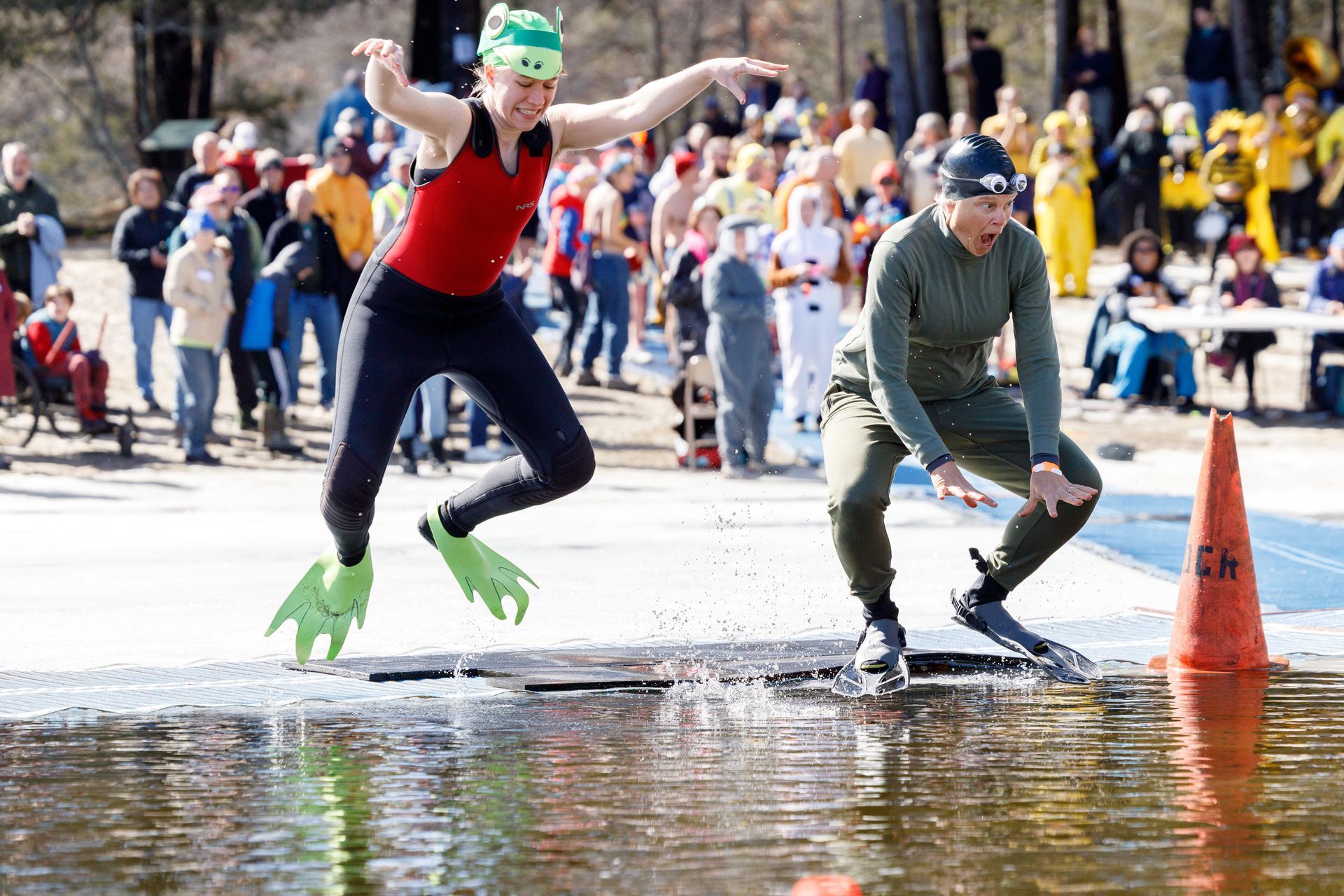Image shows 2 women, dressed as frogs, frog-hopping into water surrounded by ice. A large crowd is in the background.