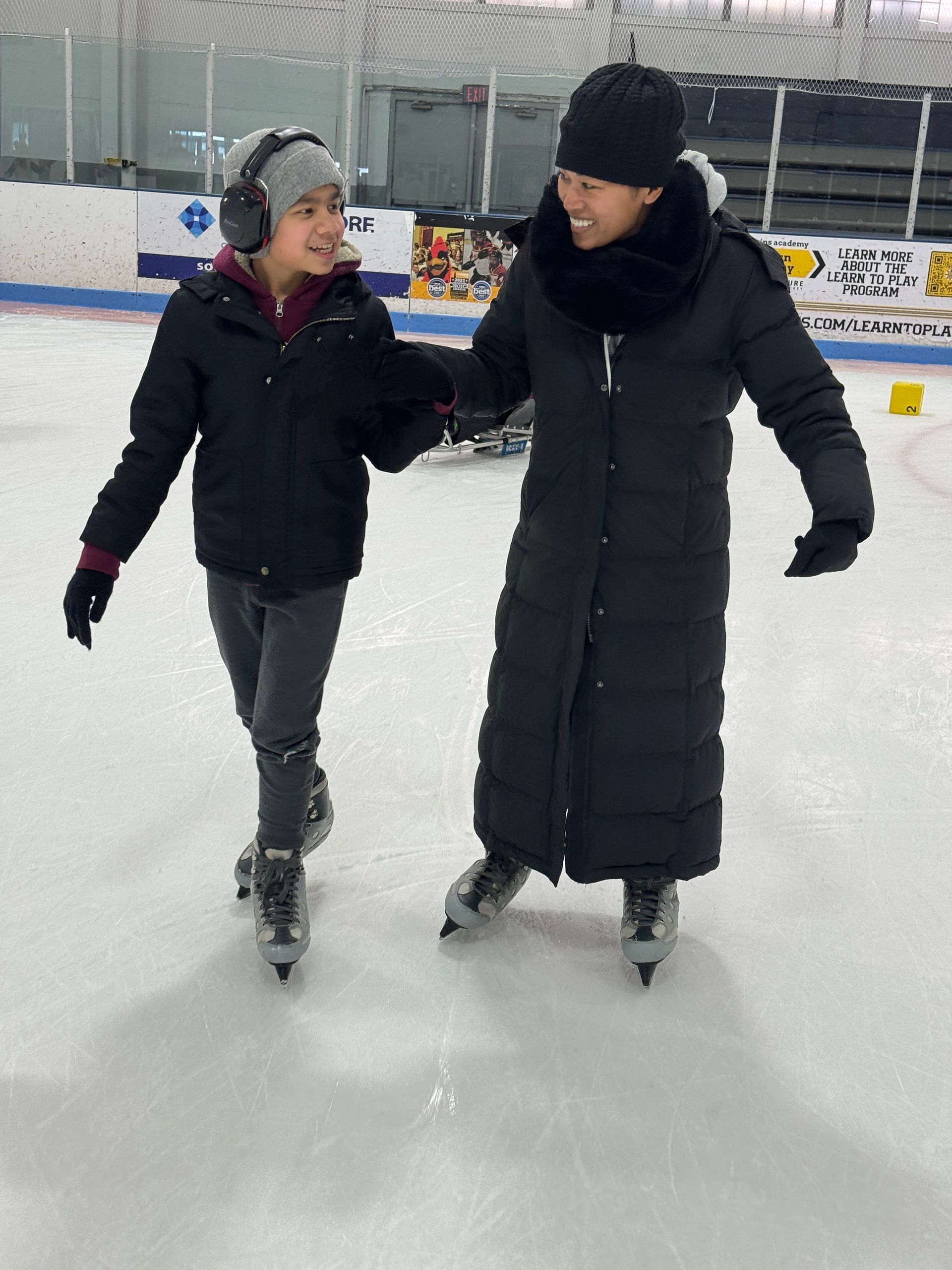 A teenager and a woman, hold hands while ice skating. They are looking at each other and smiling; the teenager is wearing noise canceling headphones.