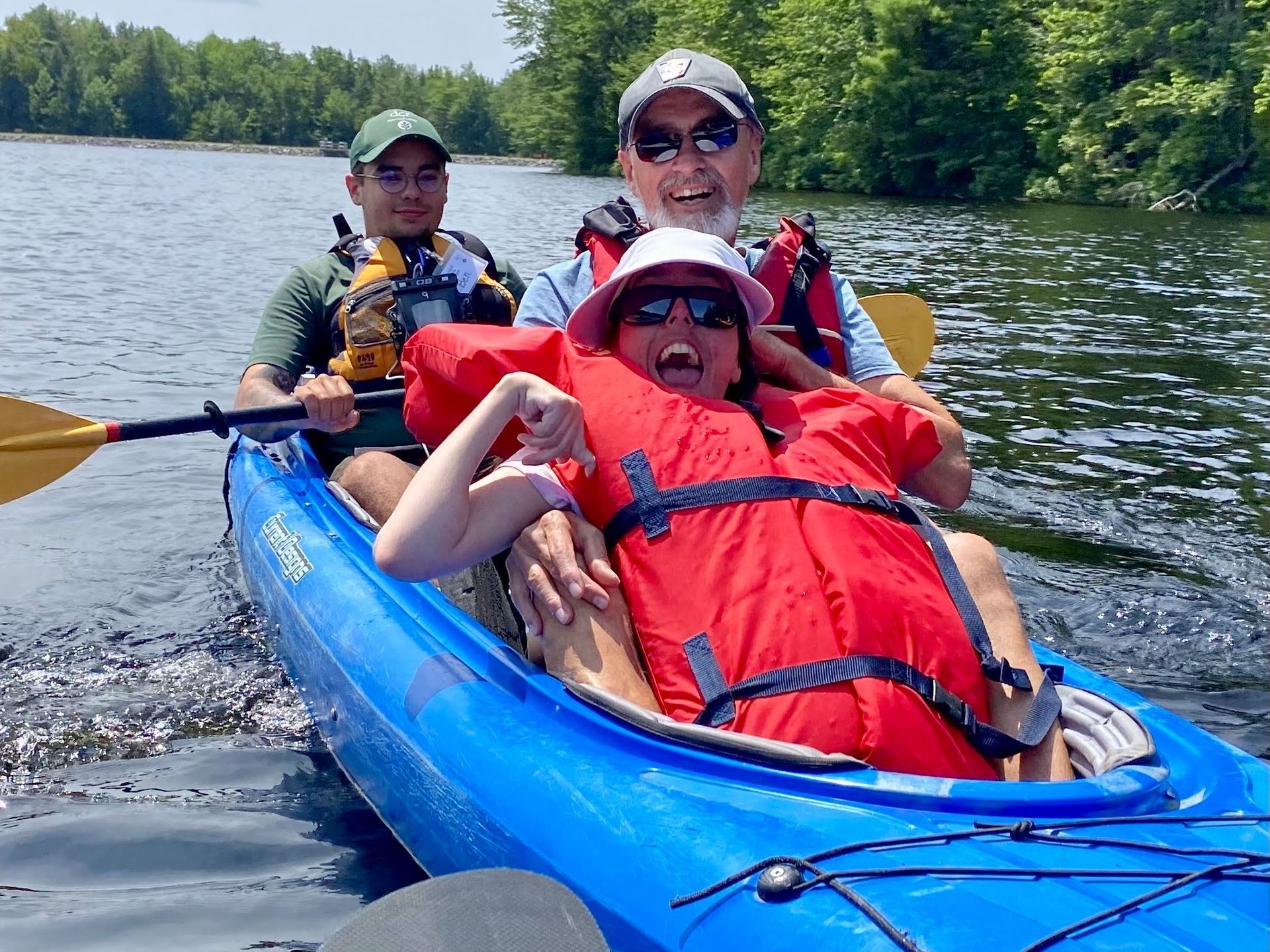 A woman in a red life lacket leans against her father in the front of a blue kayak. Both have huge smiles. Behind them, a person is holding a kayak paddle, ready to paddle their kayak.