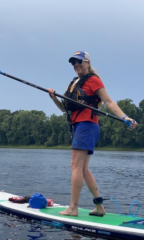 A woman in hat and sunglasses is smiling and standing on a paddle board in the water holding a paddle.