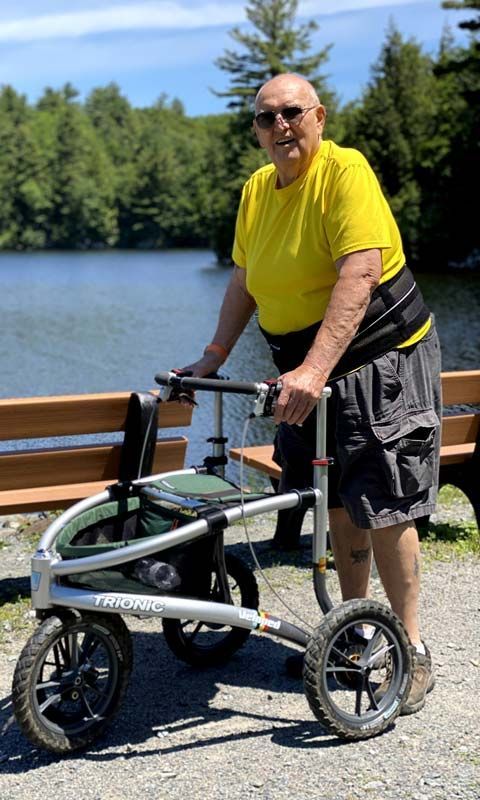 An elderly man in shorts and a t-chirt uses a walker on a hiking trail near a river.