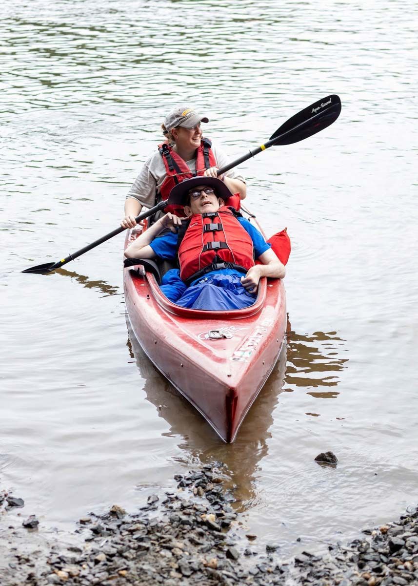 An able-bodied person paddles with a disabled passenger in a kayak near the shore of the Connecticut river.