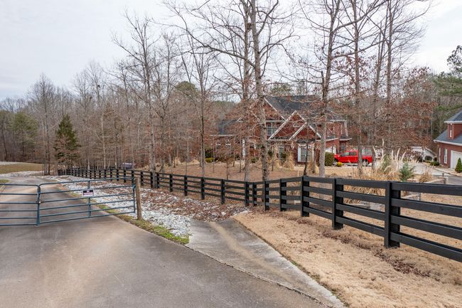 A rural driveway leads to a large, multi-story brick house behind a black wooden fence and bare trees on a cloudy day.