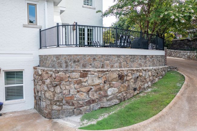 A stone retaining wall curves around the base of a white house with an elevated metal-railed deck and a driveway.