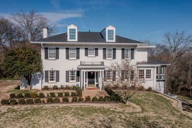 A two-story, white Colonial-style home with black shutters, dormer windows, and a front porch under a clear blue sky.