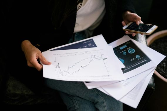 A person holding printed financial charts and graphs while looking at a smartphone screen.
