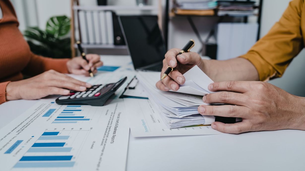 Two people sit at a table reviewing financial reports and US currency with a calculator and laptop in an office setting.