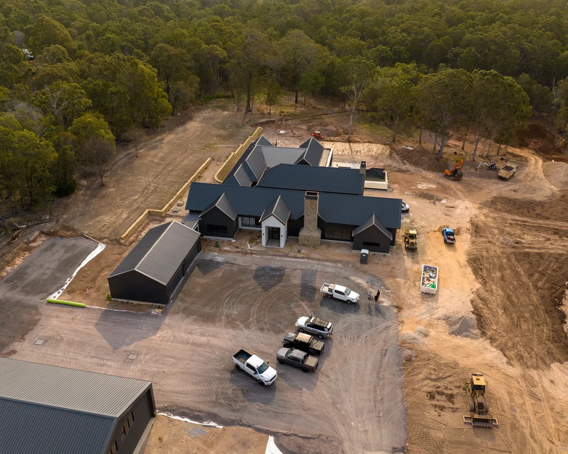 Aerial view of a dark-roofed house under construction with parked trucks and machinery in a wooded area. — Smart Metal Roofing in Coomera, QLD